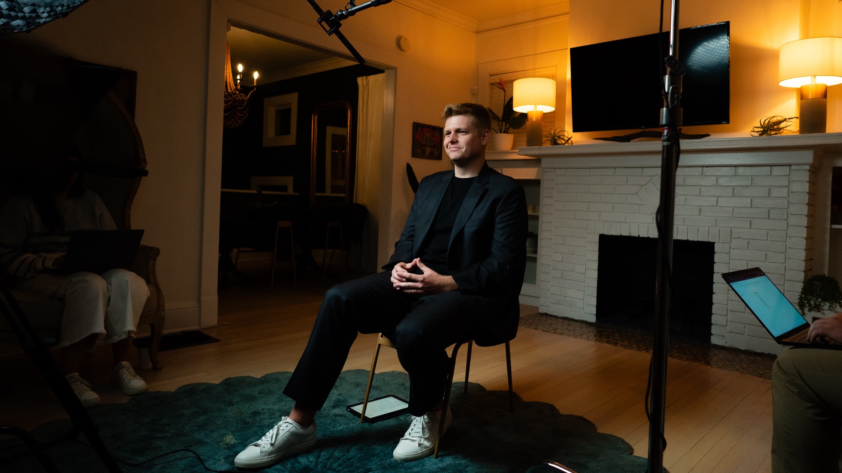 Jacob Warwick seated for an on-location interview beside a white brick fireplace, lit warm from camera right.