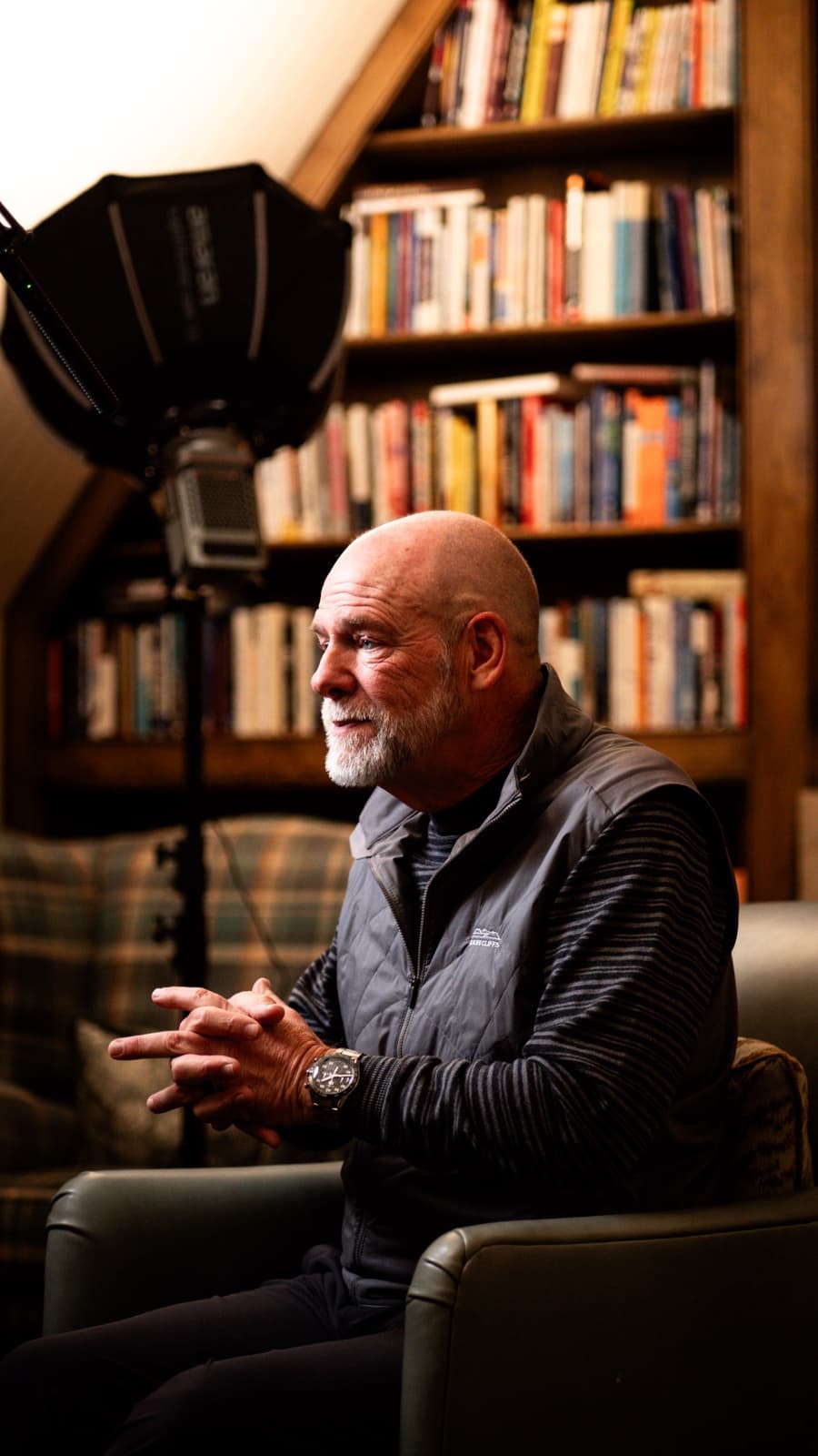 Mark Miller talking in an armchair in a book-lined attic room, a softbox rigged to camera left.